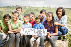 Multi-ethnic and mixed aged group of children holding a white, sign outdoors that reads "Teamwork". The group is of Latin, Asian, and Indian descent. They are sitting outdoors with a view of their countryside behind them. Collaboration, volunteerism themes.